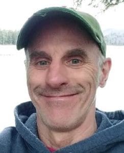 Man wearing green cap and dark blue jacket smiles at camera with lake and trees in background.