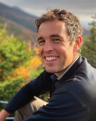 Man smiling at camera outdoors with forested mountains and green vegetation in background.