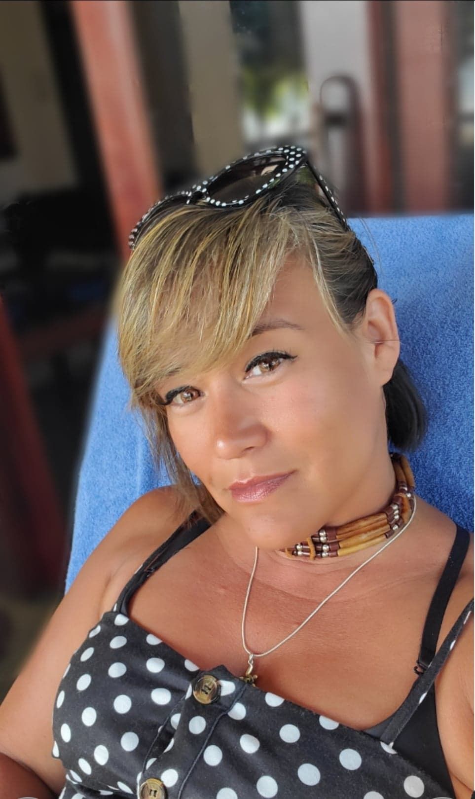 Tania Margate, with short blonde hair, wears a black and white polka dot top, sunglasses on her head, a wooden bead necklace, and a pendant necklace while seated and looking at the camera.