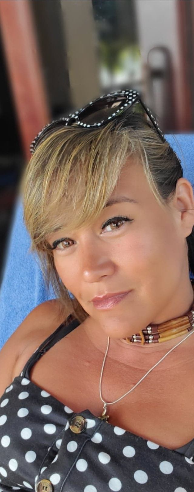 Tania Margate, with short blonde hair, wears a black and white polka dot top, sunglasses on her head, a wooden bead necklace, and a pendant necklace while seated and looking at the camera.