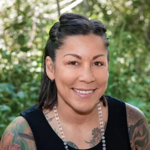 Melissa Rivera, with dark hair, wearing a sleeveless black top and beaded necklace, is smiling while sitting outdoors in a lush, green, sunlit area.