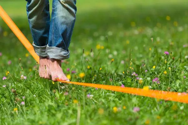 Person in jeans and bare feet walking along orange rope in grassy field with wildflowers.