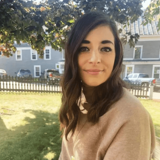 Amanda Kurlanski, in a beige sweater, sits outside on a grassy lawn with long brown hair flowing, embodying the spirit of Trusting the Process amid trees and parked cars on a sunny day.