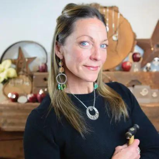 A woman with long blonde hair, wearing a black top, large earrings, and a silver necklace, stands indoors holding a hammer. Jewelry and wooden decorations fill the background, reflecting Willa Wirths Force of Nature style.