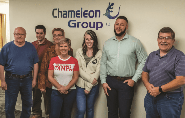 Seven people stand smiling in a row in front of the Chameleon Group LLC logo. They appear to be colleagues or a team, casually dressed, posing for a group photo at their Recovery Friendly Workplace.