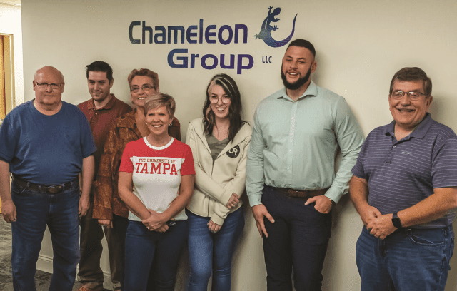 Seven people stand smiling in a row in front of the Chameleon Group LLC logo. They appear to be colleagues or a team, casually dressed, posing for a group photo at their Recovery Friendly Workplace.