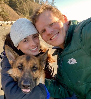 A smiling couple poses outdoors with a German Shepherd. The woman wears a white knit hat and the man, Christopher Poulos, is in a green jacket. Trees and sunlight are visible in the background.