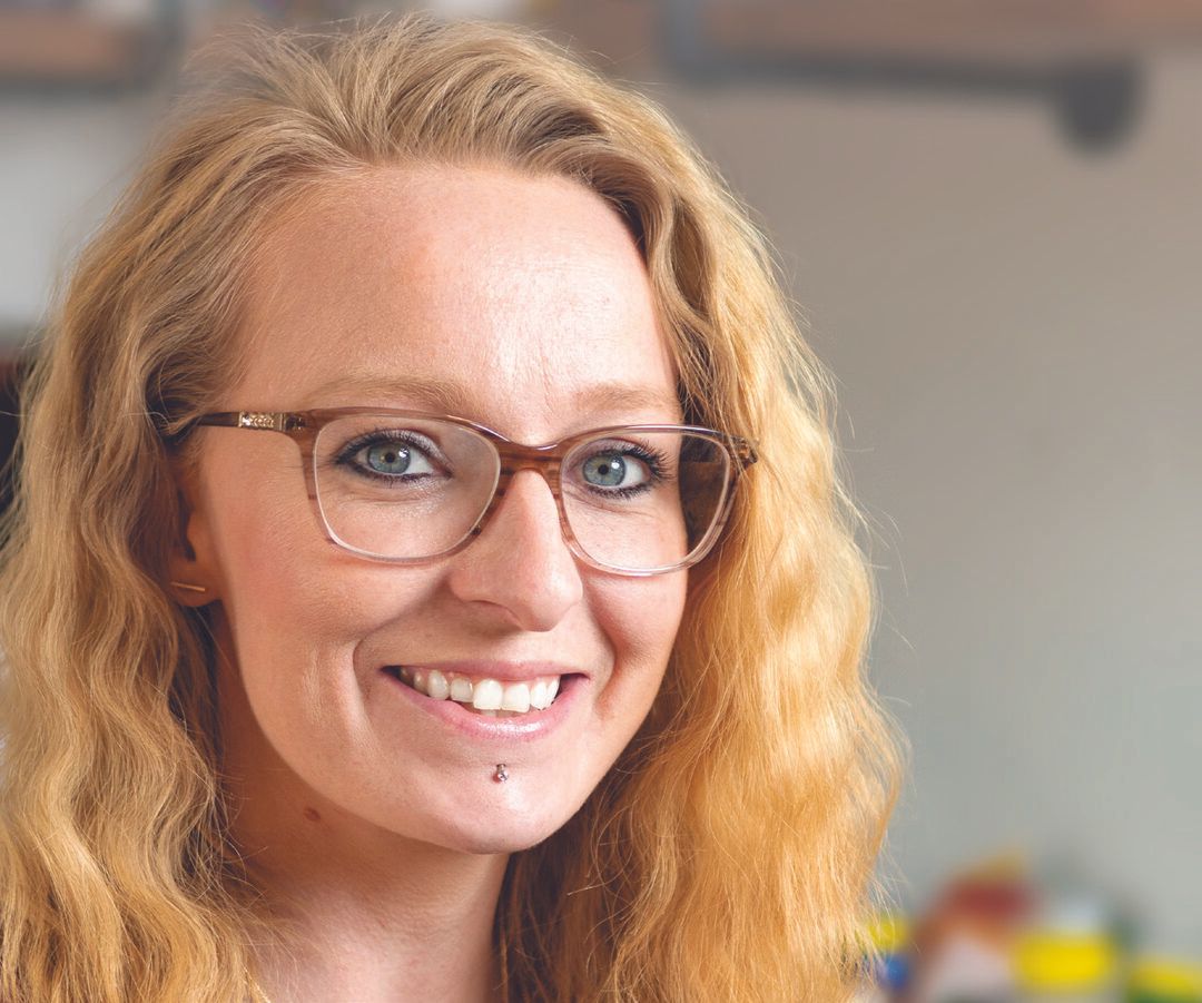 Sarah Langevin, a woman with long wavy blonde hair and glasses, smiles at the camera. She has a lip piercing and is in a brightly lit indoor setting.