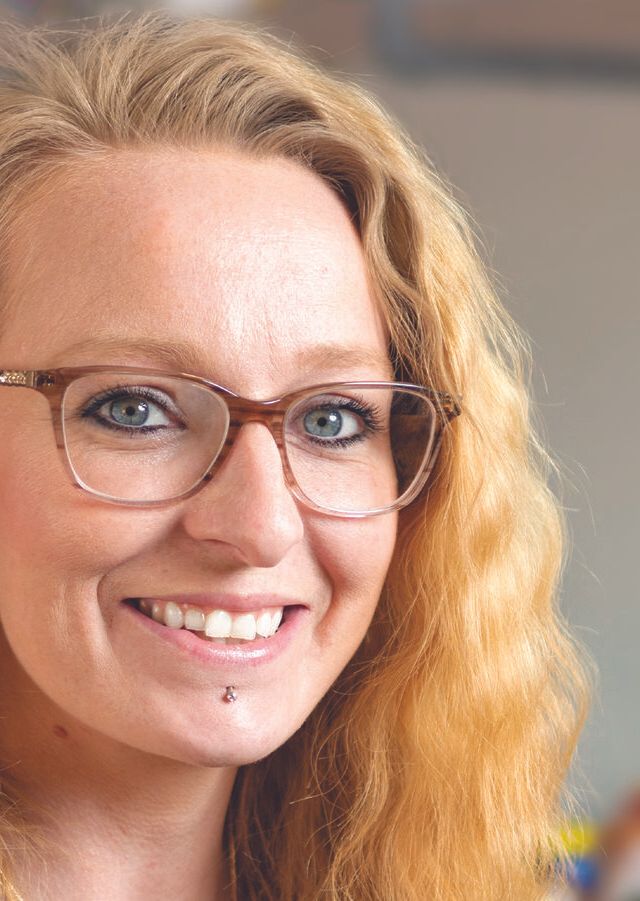 Sarah Langevin, a woman with long wavy blonde hair and glasses, smiles at the camera. She has a lip piercing and is in a brightly lit indoor setting.