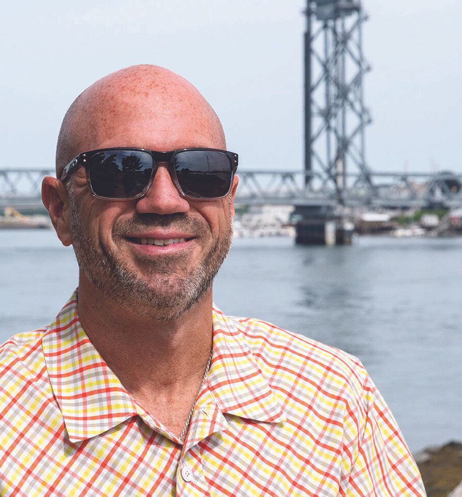 Freddy Petrone, a bald man wearing sunglasses and a yellow plaid shirt, smiles while standing near a body of water, with a bridge in the background on a bright day.