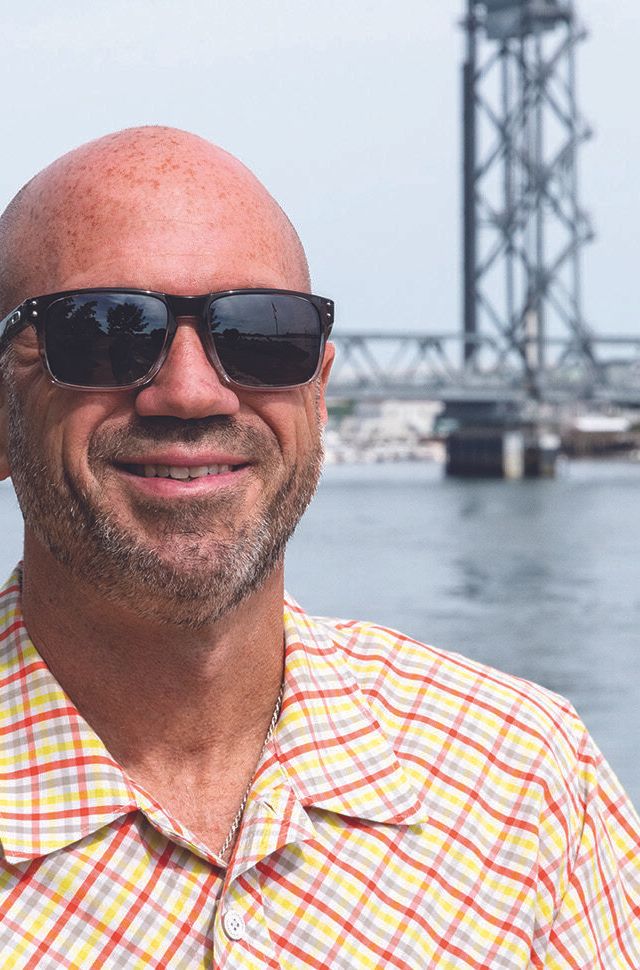 Freddy Petrone, a bald man wearing sunglasses and a yellow plaid shirt, smiles while standing near a body of water, with a bridge in the background on a bright day.