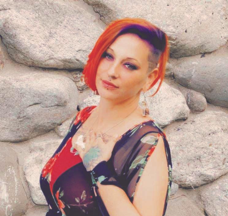 Christine Maider, with vibrant red and purple hair, wearing a floral dress and earrings, stands before a wall of large gray stones. She poses with one hand on her chest, looking at the camera. The image appears pixelated.