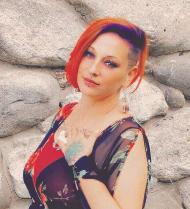 Christine Maider, with vibrant red and purple hair, wearing a floral dress and earrings, stands before a wall of large gray stones. She poses with one hand on her chest, looking at the camera. The image appears pixelated.