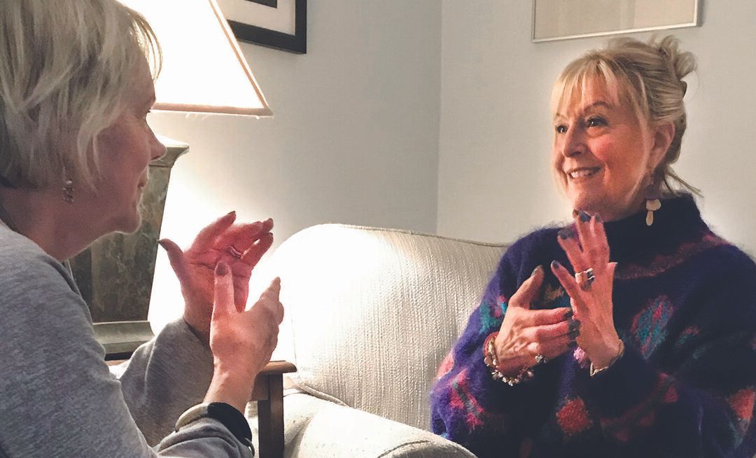 Two women sit facing each other on a couch, smiling and gesturing with their hands as they have a lively conversation about self-care in a cozy, warmly lit room.