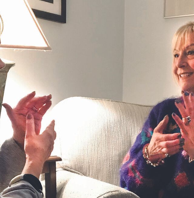 Two women sit facing each other on a couch, smiling and gesturing with their hands as they have a lively conversation about self-care in a cozy, warmly lit room.