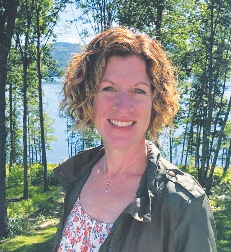A woman with curly, short hair smiles outdoors in Maine, wearing a floral top and green jacket. Behind her are tall trees, green foliage, and a lake with hills in the background—a bright day captured by Maine Voices of Recovery.