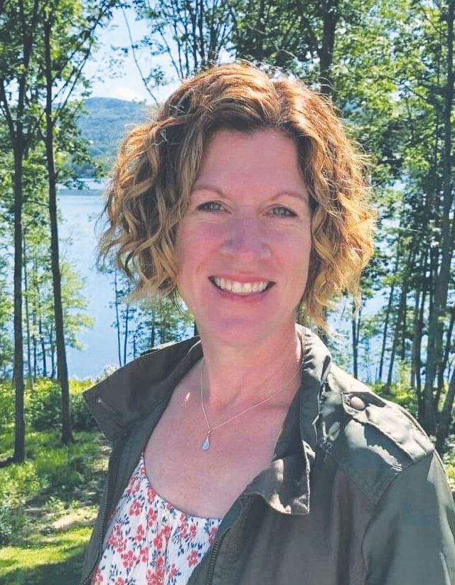 A woman with curly, short hair smiles outdoors in Maine, wearing a floral top and green jacket. Behind her are tall trees, green foliage, and a lake with hills in the background—a bright day captured by Maine Voices of Recovery.