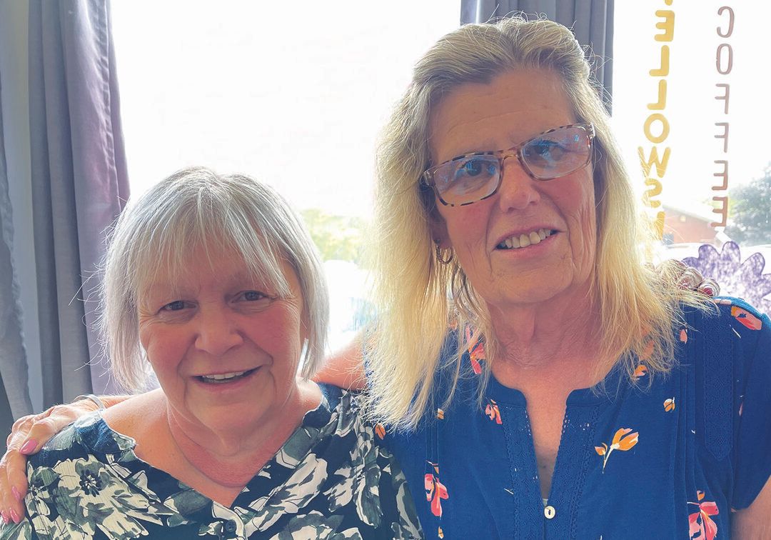 Two older women smile together indoors at the Recovery Community Center, one with short gray hair in a floral top, the other with long blonde hair, glasses, and a blue shirt. Sunlight streams through the window behind them.