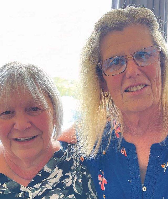 Two older women smile together indoors at the Recovery Community Center, one with short gray hair in a floral top, the other with long blonde hair, glasses, and a blue shirt. Sunlight streams through the window behind them.