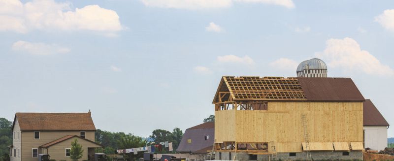 A partially constructed barn with exposed wooden framework stands beside a traditional farmhouse and silo under a partly cloudy sky—an image reflecting the strength of rural communities and the spirit of coalition building.