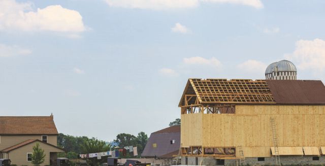 A partially constructed barn with exposed wooden framework stands beside a traditional farmhouse and silo under a partly cloudy sky—an image reflecting the strength of rural communities and the spirit of coalition building.