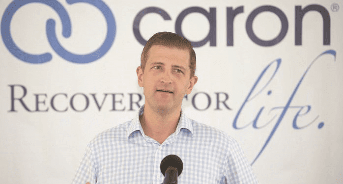 A man in a checked shirt speaks at a microphone in front of a Caron banner with the slogan Recovery for life.