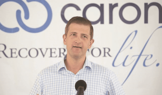 A man in a checked shirt speaks at a microphone in front of a Caron banner with the slogan Recovery for life.