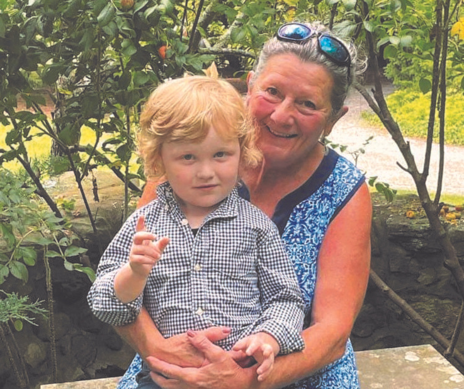 An older woman in a blue dress smiles while hugging a young child with curly blond hair, both enjoying the outdoors in a green garden setting, embodying the warmth and connection celebrated by Maine Voices of Recovery.