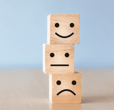 Three wooden blocks are stacked on top of each other, each featuring a different face: a sad face on the bottom, a neutral in the middle, and a happy face on top—like an emotional Sensational Radar against a blue background.