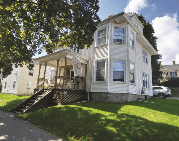A two-story beige Sober Living House with white trim, a covered front porch, and large windows sits on a grassy lawn under a tree, offering a Fresh Start beneath blue skies with a car parked in the driveway.