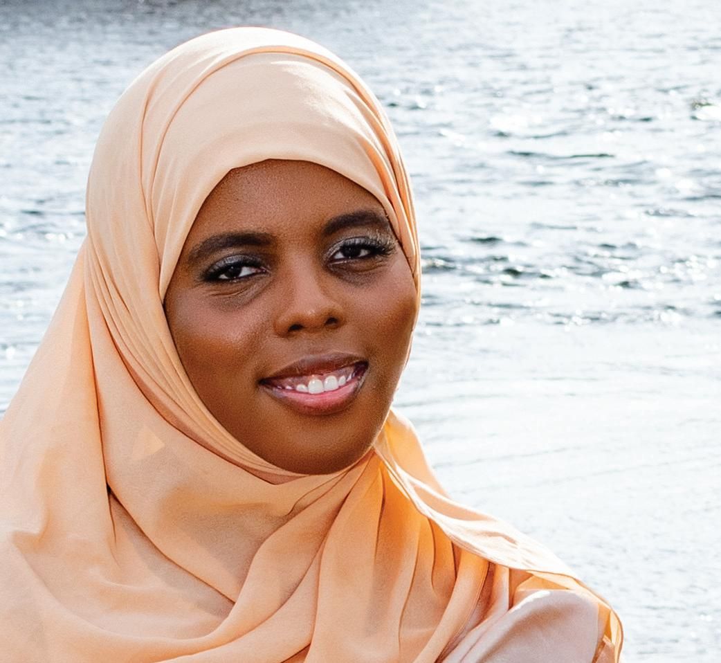 A woman wearing a light peach hijab, embodying Generational Noor, smiles gently while standing outdoors near a body of water.
