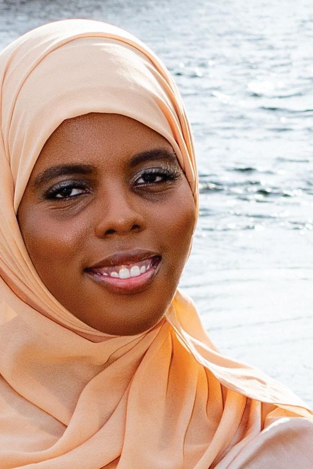 A woman wearing a light peach hijab, embodying Generational Noor, smiles gently while standing outdoors near a body of water.