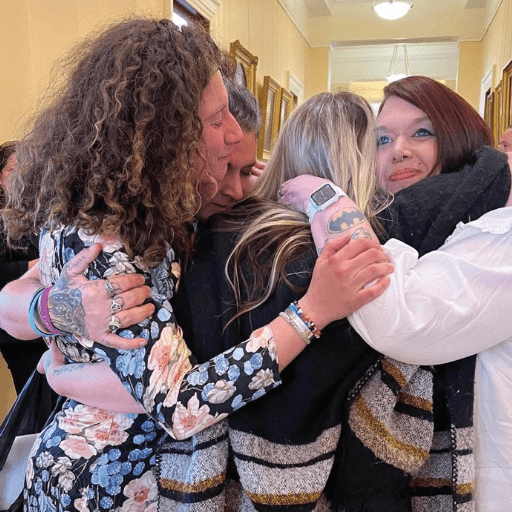 Four people stand in a group hug in a hallway with framed pictures on the wall. Two have visible tattoos, and all appear emotional and supportive. Warm lighting fills the scene, echoing themes of compassion and knowing your rights in Maine.