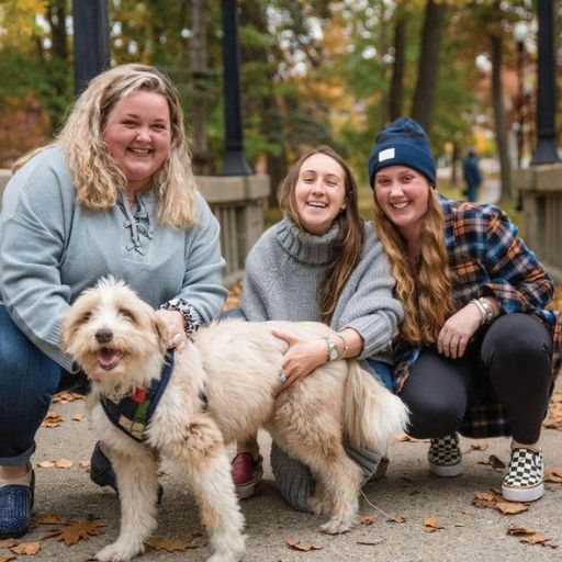 Three smiling women, including Carly Flagg, pose together outdoors on a leaf-covered path in autumn, crouching next to a happy, fluffy light-colored dog. Trees with fall foliage and a bridge appear in the background.