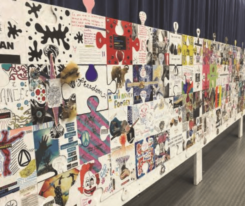 A large display of colorful, decorated puzzle pieces forms a mural, each piece featuring different drawings, photos, words, and designs—creating a ripple effect—standing on a white frame in front of a dark curtain.