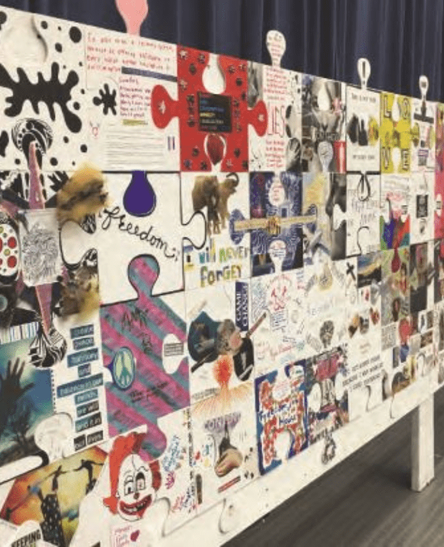 A large display of colorful, decorated puzzle pieces forms a mural, each piece featuring different drawings, photos, words, and designs—creating a ripple effect—standing on a white frame in front of a dark curtain.