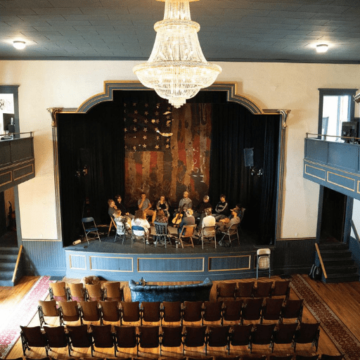 A group of people with open hearts sits in a circle onstage under a large chandelier in a small vintage theater with an American flag backdrop; empty chairs fill the audience area, echoing the hopes of small-town filmmakers to change perceptions.