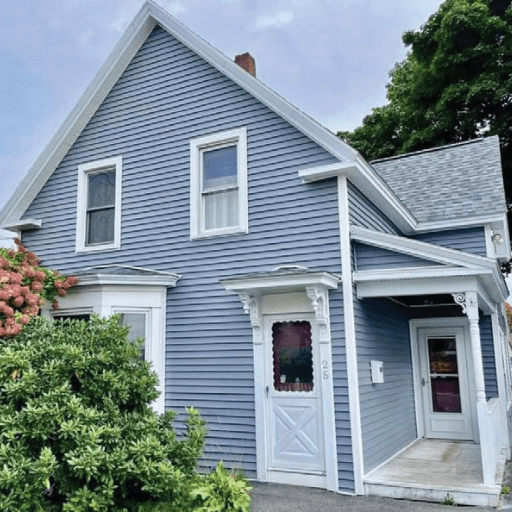 A blue two-story house with white trim sits in New Hope Alliance, featuring a covered porch and a red door. Green bushes and flowering plants line the front, while large trees provide a peaceful background.