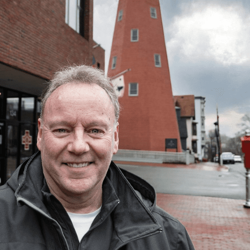 A smiling man wearing a jacket stands on a sidewalk in front of a brick building with a tall, pointed tower on a cloudy day, capturing the spirit of an Ed Crockett story.