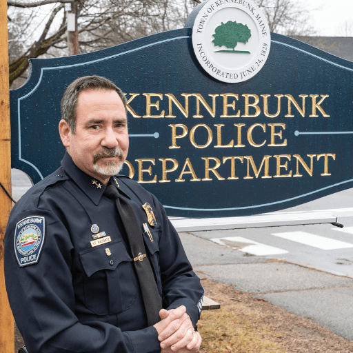 A police officer in uniform stands in front of a sign that reads Kennebunk Police Department, embodying the dedication of Law Enforcement Officers, with the town seal of Kennebunk, Maine visible at the top.
