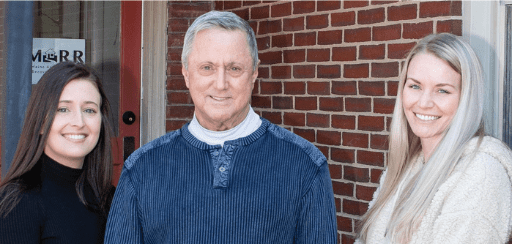 Three people, two women and one man, are standing outside in front of a brick wall, smiling at the camera. The man, in a blue sweater, stands between the two women. They represent the Maine Association of Recovery Residences.