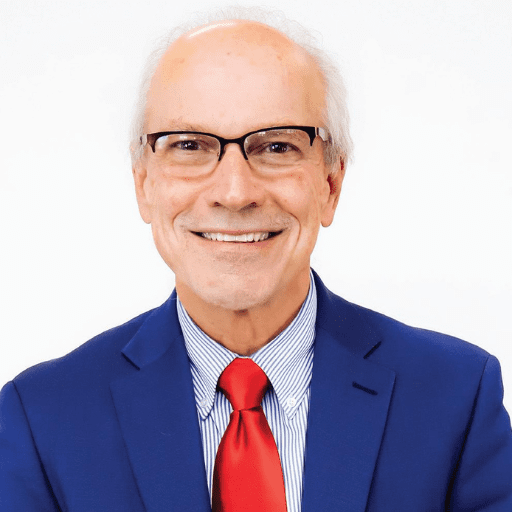 Gordon Smith, a smiling older man with glasses, short white hair, and wearing a blue suit jacket, white and blue striped shirt, and red tie, poses in front of a plain white background.