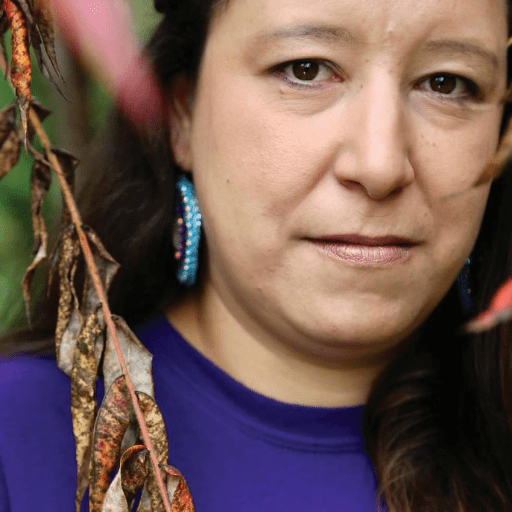 April Tomah, with long dark hair and beaded earrings, stands outdoors beside dried leaves. Wearing a purple top, she looks directly at the camera with a neutral expression, embodying strength and the spirit of someone who has recovered.