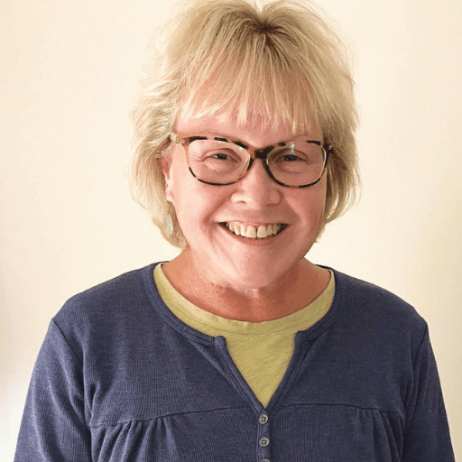 A smiling woman with short blonde hair and glasses, wearing a blue top over a light yellow shirt, stands in front of a plain light-colored background, radiating the confidence and positivity that come with the benefits of sobriety.