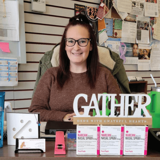 A woman with glasses and a brown sweater sits smiling at a desk with a Gather sign, Narcan nasal spray boxes, and office supplies. A bulletin board glows under bright light at the Washington County Centers in the background.