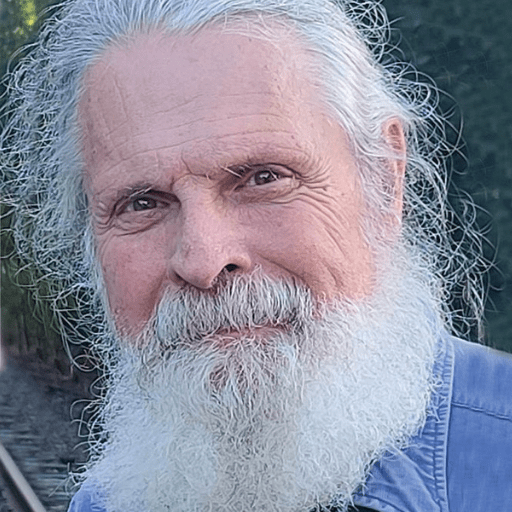 Jim Mello, known for Stories Restoring Hope, is an older man with a long white beard and hair, wearing a blue shirt. He stands outdoors near railroad tracks with greenery in the background, looking into the camera and smiling gently.