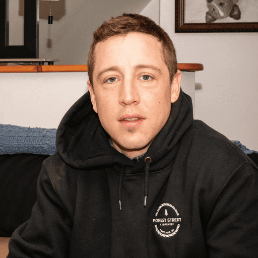 A person with short light brown hair wearing a black hoodie with a Forest Street Carpentry logo sits indoors, looking at the camera. Building Better, they embody craftsmanship amid a cozy setting with a couch and framed artwork in the background.