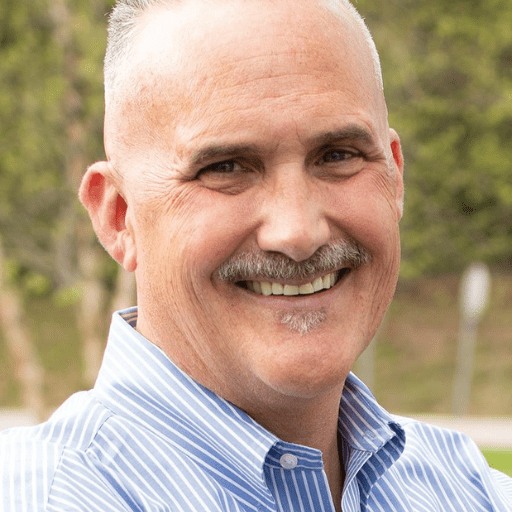 A middle-aged man with short gray hair and a mustache smiles outdoors, wearing a light blue collared shirt. The background is blurred greenery.