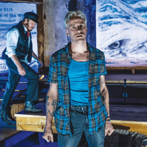 A young man with blond hair, tattoos, and a blue plaid shirt stands center stage. Another man in period clothing and a hat walks behind him. The nautical scene, titled Ahab Inside Me, features a ship’s wheel and painted wave backdrop.