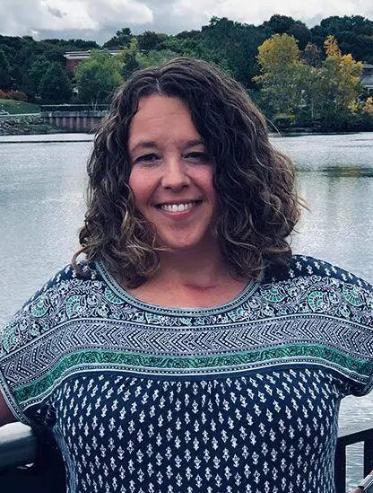A woman with curly brown hair smiles at the camera while standing in front of a lake, embodying what it means to be a Recovery Ally—showing that no special skills are needed to support others. Trees and a cloudy sky form the background as she wears a blue and green top.